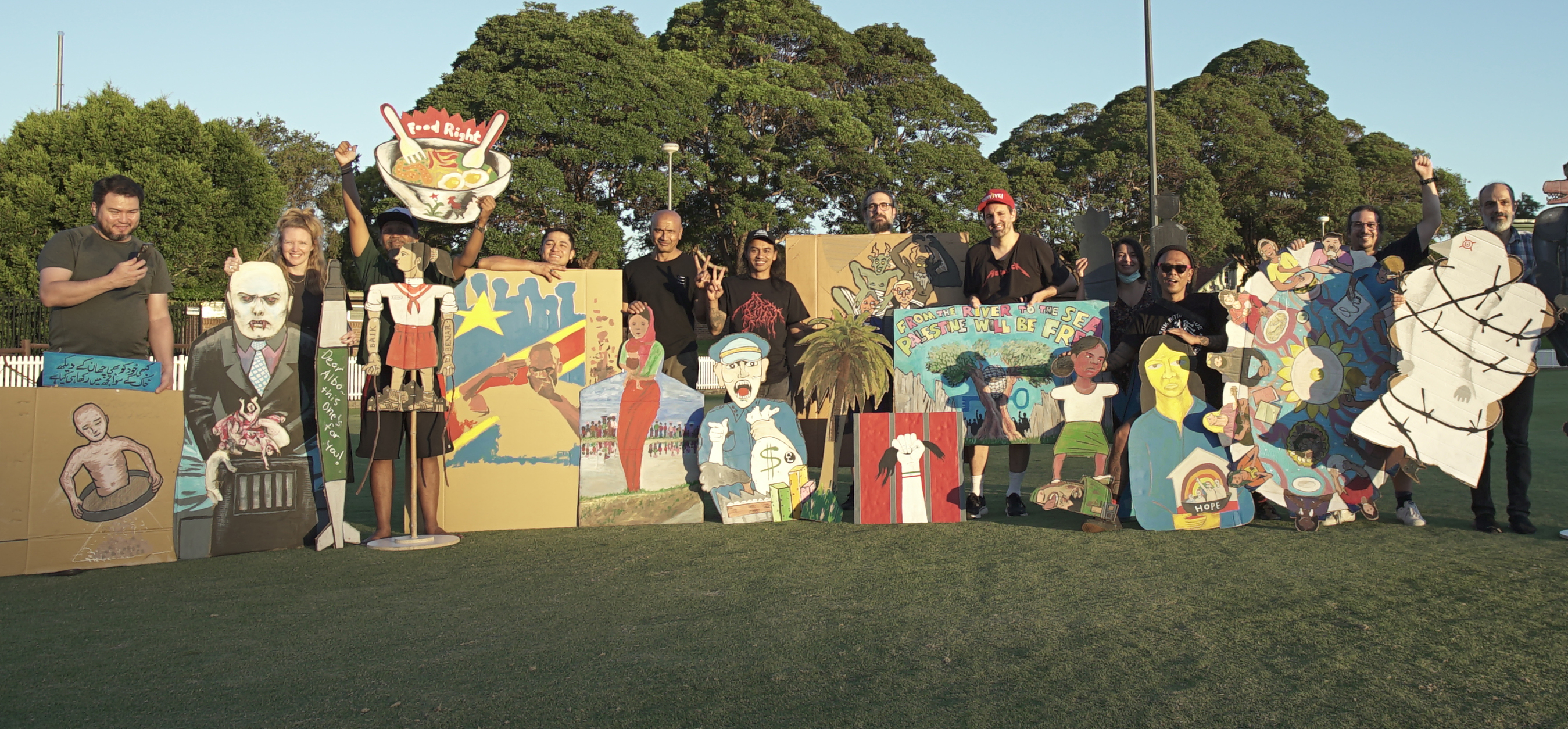 Participating artists stand with their creations, mostly large cardboard cut out painted artworks inspired by the Indonesian puppet style made by Taring Padi, the artists stand on Pratten Park oval with the sun shining on them, there are about 14 artists with colourful political artworks